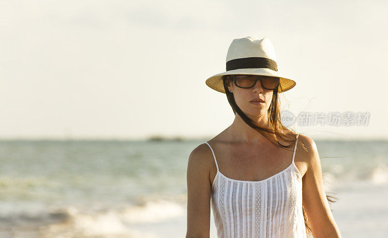 美丽的女人穿着白色的裙子，太阳镜和草帽走在田诗般的São米格尔dos Milagres海滩在日落，阿拉格斯，巴西东北部